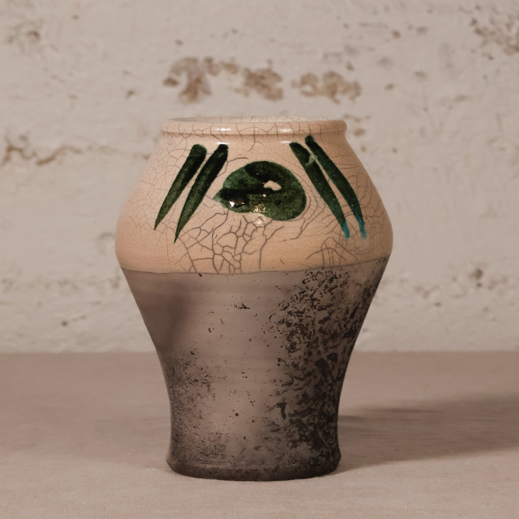 A handmade ceramic vase with a textured, crackled finish, featuring a beige upper half with green painted abstract stripes and a dark gray, speckled lower half, set against a rustic, light-colored textured wall.