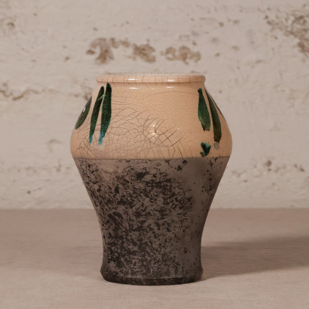 A handmade ceramic vase with a two-tone finish, featuring a warm beige upper half with a crackled glaze and green painted leaf motifs, and a dark gray speckled lower half, set against a rustic textured wall.