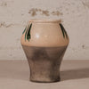 A handmade ceramic vase with a cream-colored crackled glaze on the upper half, decorated with green leaf motifs, and a textured dark gray-brown lower half, set against a rustic plaster wall.
