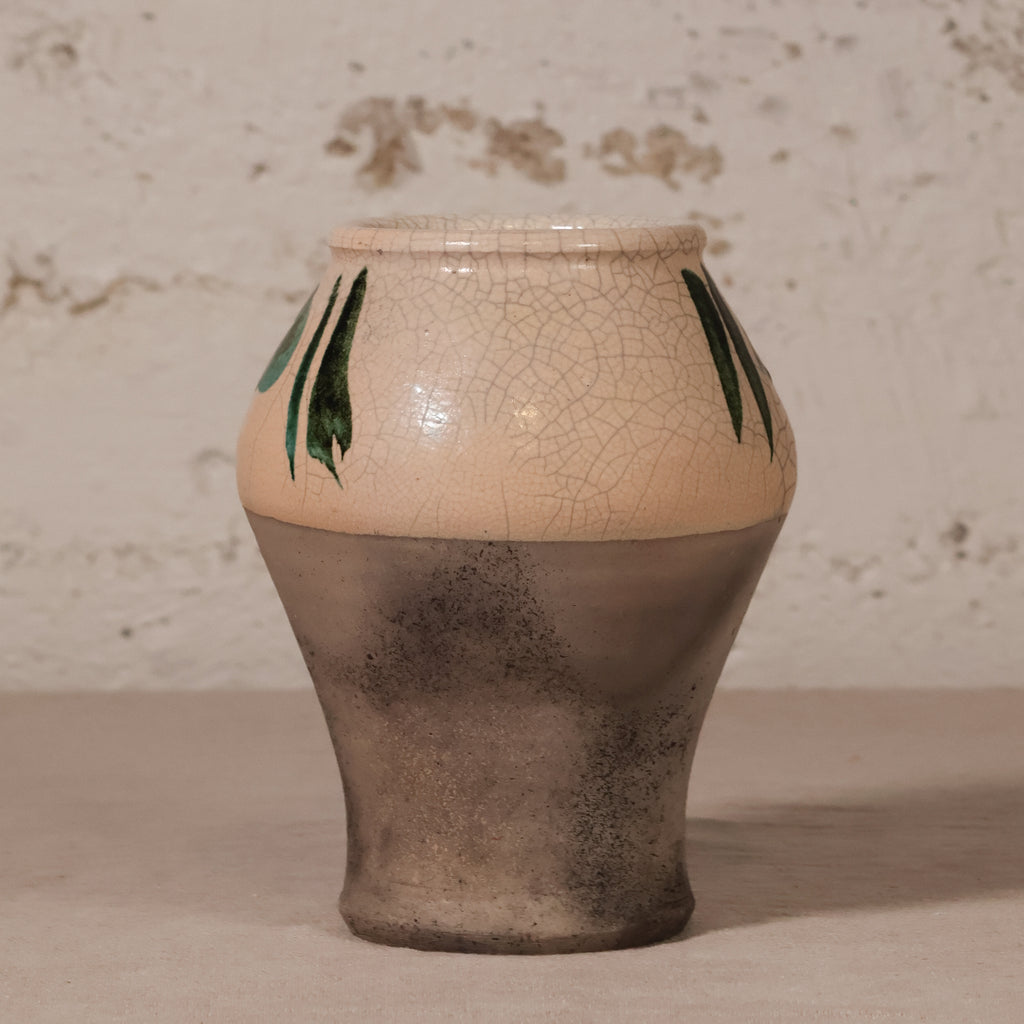 A handmade ceramic vase with a cream-colored crackled glaze on the upper half, decorated with green leaf motifs, and a textured dark gray-brown lower half, set against a rustic plaster wall.
