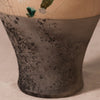 Close-up of a ceramic vase or vessel with a mottled dark gray to black lower body and a warm beige upper section with visible crackle glaze and small green glaze accents.
