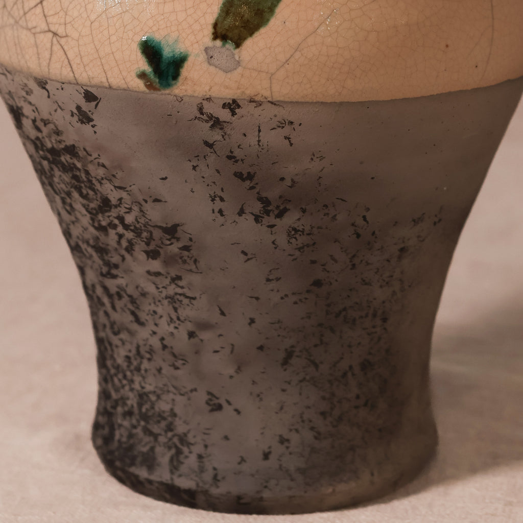 Close-up of a ceramic vase or vessel with a mottled dark gray to black lower body and a warm beige upper section with visible crackle glaze and small green glaze accents.
