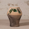 A handmade ceramic vase with a two-tone design, featuring a warm beige upper half decorated with green leaf patterns and a textured dark gray-brown lower half, set against a rustic white plaster wall.