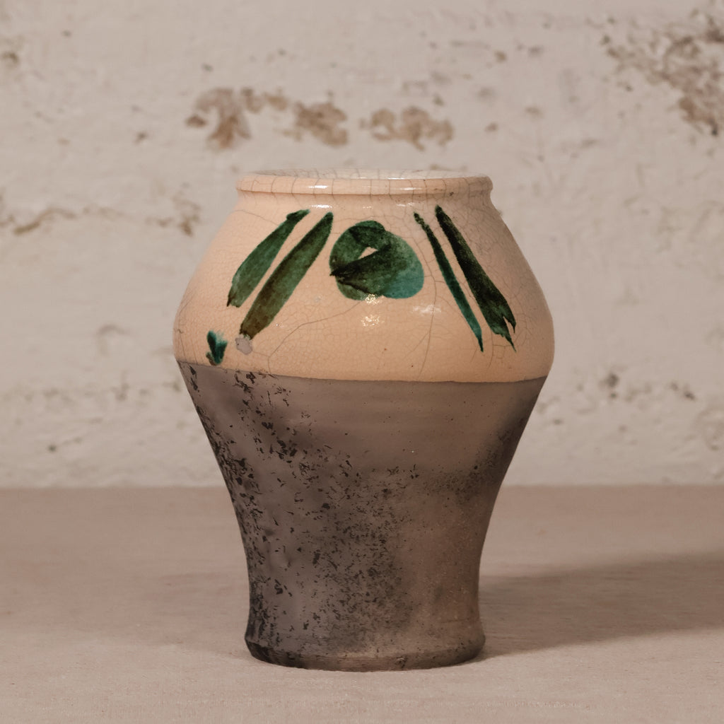 A handmade ceramic vase with a two-tone design, featuring a warm beige upper half decorated with green leaf patterns and a textured dark gray-brown lower half, set against a rustic white plaster wall.