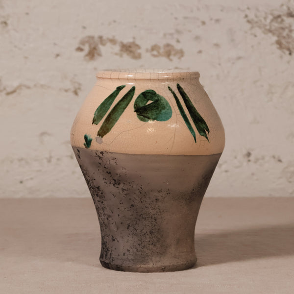 A handmade ceramic vase with a two-tone design, featuring a warm beige upper half decorated with green leaf patterns and a textured dark gray-brown lower half, set against a rustic white plaster wall.