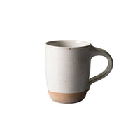 A handmade ceramic mug with a matte off-white body, a textured light brown base, and a curved handle, set against a neutral gray surface and a textured light green wall.
