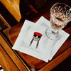 A close-up of a white paper napkin featuring a cartoon illustration of a person with braids, sunglasses, and a red beret, placed on a polished wooden surface next to a clear stemmed glass.