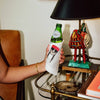 A hand holding a green glass bottle wrapped in a white sleeve printed with a black-and-white illustration of a skull wearing a red hat. The scene is indoors, with a black lamp, decorative figurine, and table visible in the background.