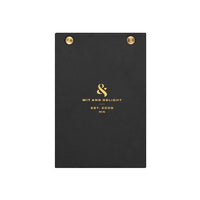 A black textured clipboard with gold metal grommets at the top and gold foil branding for 'Wit and Delight' in the center, shown on a plain white background.