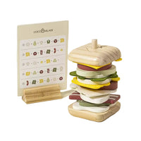 A wooden toy sandwich stack by Coco Village on a light beige background, featuring a multi-layered wooden burger with yellow, green, red, and white layers, next to a matching wooden instruction card and small wooden stand.